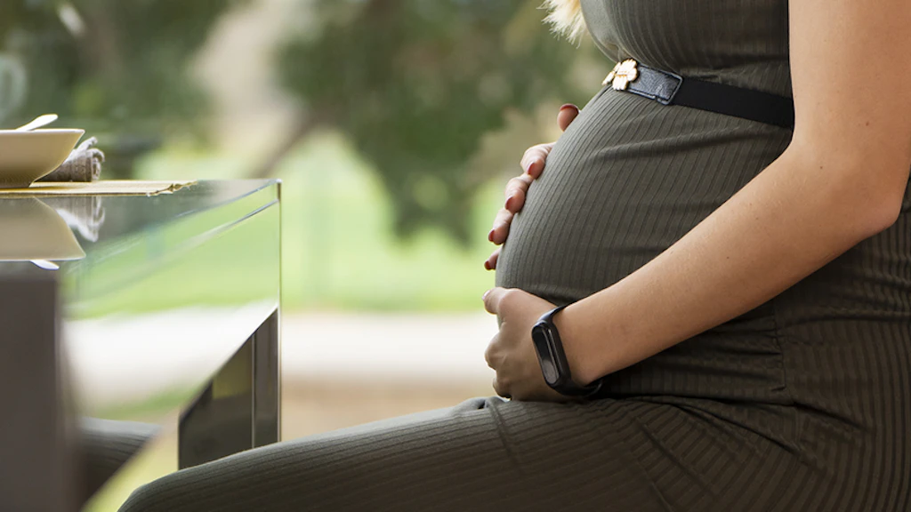 Schwangere Frau sitzt entspannt an einem Tisch, hält ihre Hände auf den Bauch.