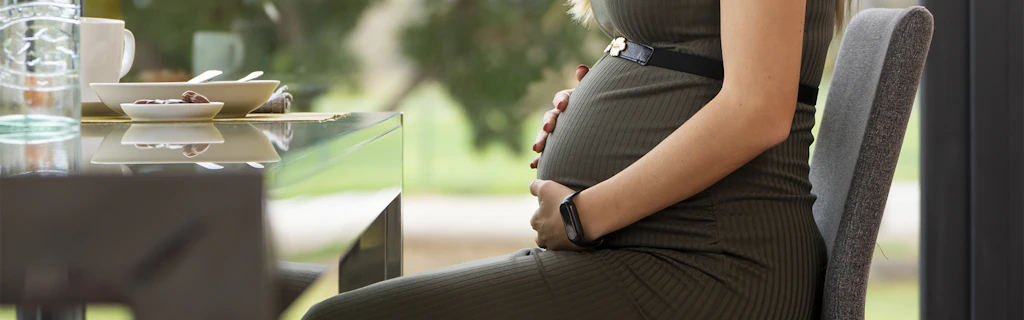 Schwangere Frau sitzt entspannt an einem Tisch, hält ihre Hände auf den Bauch.