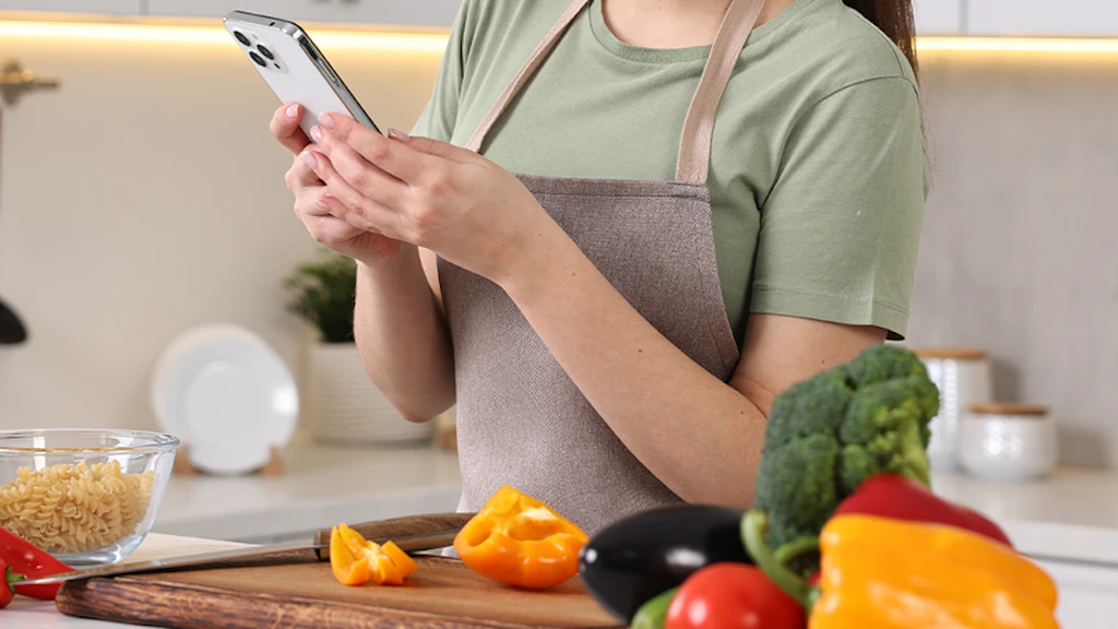 Frau, deren Kopf man nicht sieht, seht am Küchentresen und sucht etwas im Handy, dass sie in der Hand hält. Auf dem Küchentresen befinden sich verschiedene Gemüsesorten sowie eine Schale mit Fusilli.
