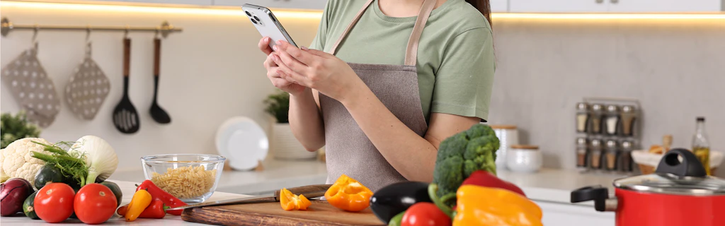 Frau, deren Kopf man nicht sieht, seht am Küchentresen und sucht etwas im Handy, dass sie in der Hand hält. Auf dem Küchentresen befinden sich verschiedene Gemüsesorten sowie eine Schale mit Fusilli.