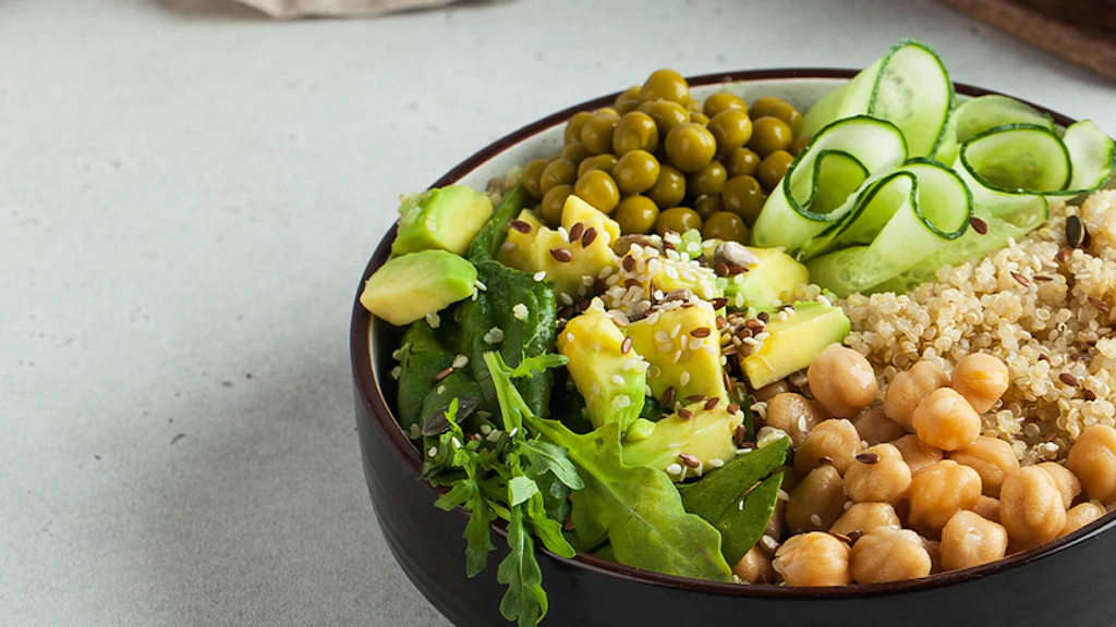 Eine Schüssel mit frischen, pflanzlichen Zutaten ist appetitlich angerichtet. Zu sehen sind unter anderem Kichererbsen, Quinoa, Salat, Gurken und weitere Gemüse.