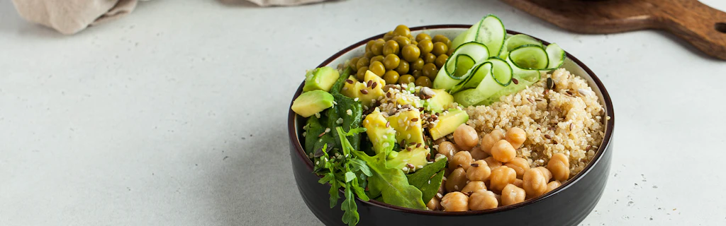 Eine Schüssel mit frischen, pflanzlichen Zutaten ist appetitlich angerichtet. Zu sehen sind unter anderem Kichererbsen, Quinoa, Salat, Gurken und weitere Gemüse.